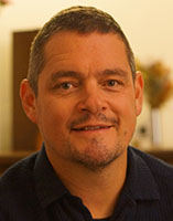A man with short brown hair and light stubble smiling at the camera.