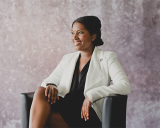 A smiling woman with dark hair wearing a white blazer and black top sits in a black chair against a textured wall.