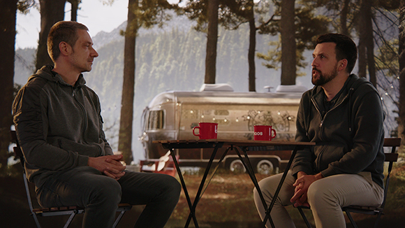 Two men sit at a small folding table in a wooded area with a silver trailer in the background.