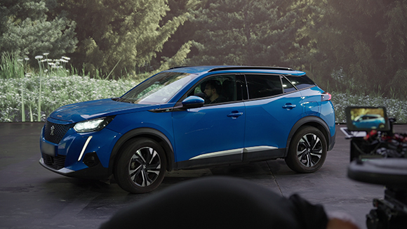 A blue Peugeot compact SUV parked in a studio with a large forest backdrop and filming equipment in the foreground.