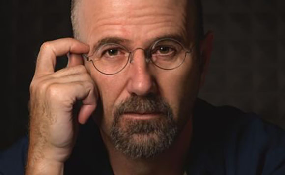A man with a salt-and-pepper beard and round glasses adjusts the frame with his hand.
