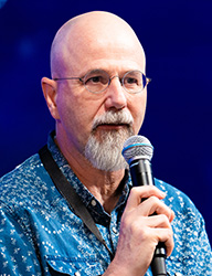 Bald man with glasses and a grey goatee speaking into a handheld microphone.