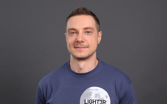 A man with short brown hair and a blue t-shirt smiling against a solid gray background.