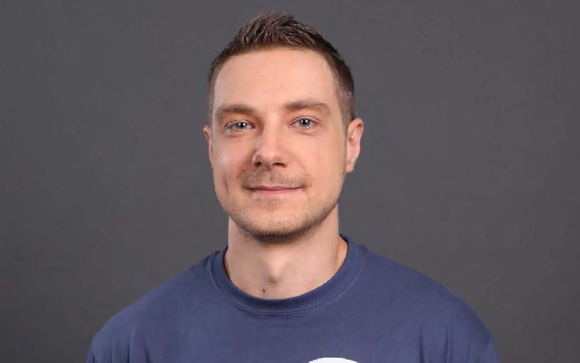 A man with short brown hair and blue eyes smiling at the camera against a solid gray background.