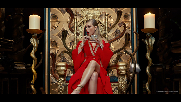 A woman in a red gown sits on a golden throne flanked by snake-shaped candle holders and lit candles.