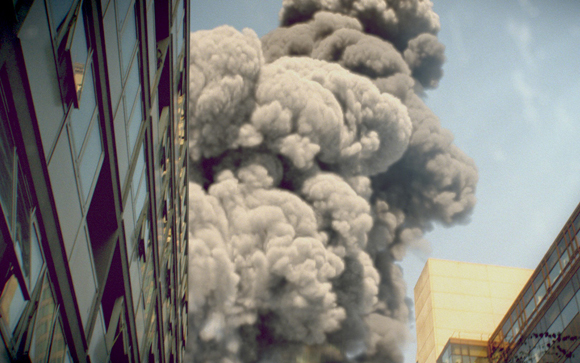 A large, billowing cloud of gray smoke rises between tall urban buildings under a clear sky.