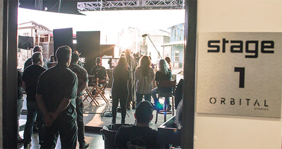 A film crew stands in a studio doorway looking toward a brightly lit set with equipment and lighting.