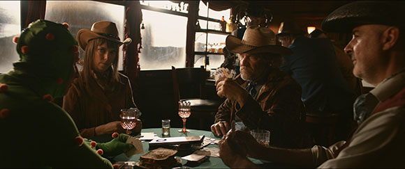 A group of people sit around a table playing cards in a dimly lit, rustic indoor setting.
