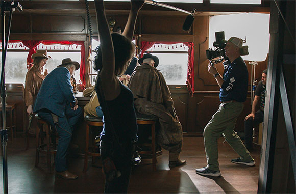 A camera operator films actors dressed in period clothing inside a wooden ship cabin.