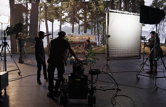 Film crew operating cameras and lighting equipment on a set with trees and a vintage trailer behind them.