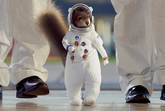 A squirrel wearing a miniature white spacesuit walks between two people in white jumpsuits.