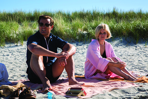 A man in a dark shirt and a woman in a pink robe sit on a pink blanket on a sandy beach.