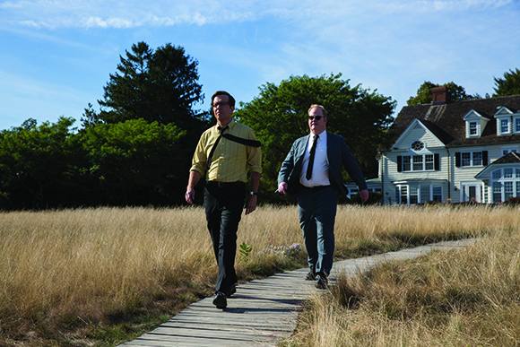Two men in business attire walk along a wooden path through a field of tall dry grass under a blue sky.