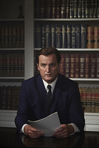 A man in a dark suit and tie sits at a desk holding a piece of paper in front of a large bookshelf.