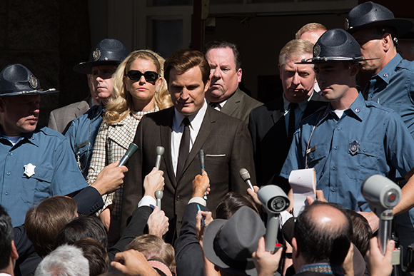 A man in a brown suit stands amidst security officers and several microphones held by unseen people.