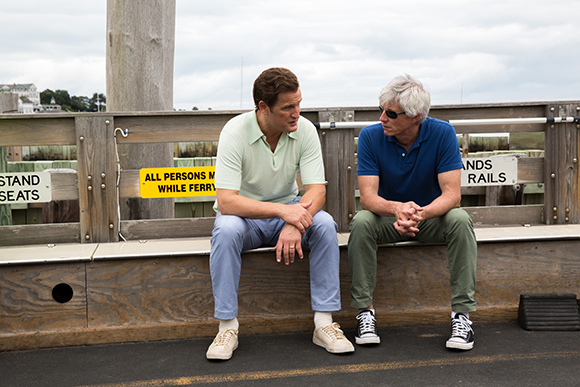 Two men sit on a wooden ledge outdoors, engaged in conversation near a ferry railing.