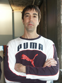 A man with dark hair stands with his arms crossed wearing a white and dark purple Puma sweatshirt.