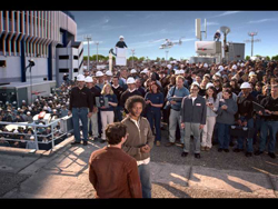 A large crowd of people standing in an outdoor plaza in front of a modern building under a blue sky.