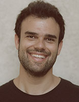 A man with dark hair and stubble smiles widely, showing his teeth. He is wearing a dark t-shirt.
