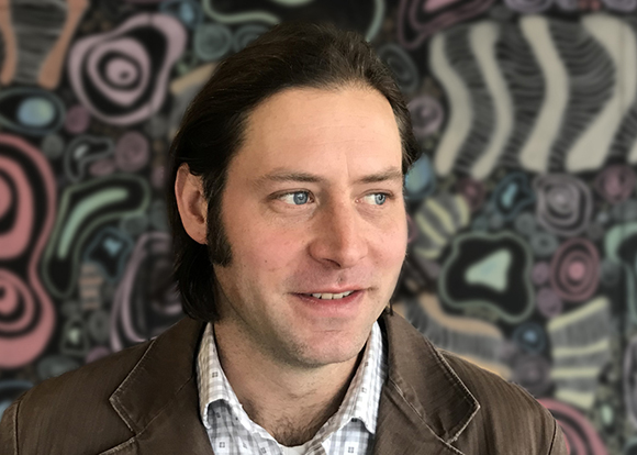 A man with dark hair and blue eyes looking off-camera against an abstract, colorful patterned backdrop.