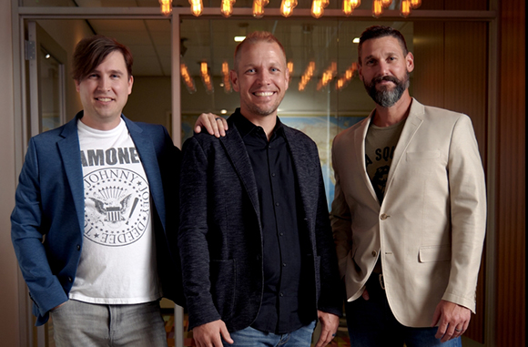Three men smiling and posing together, wearing blazers and casual shirts.