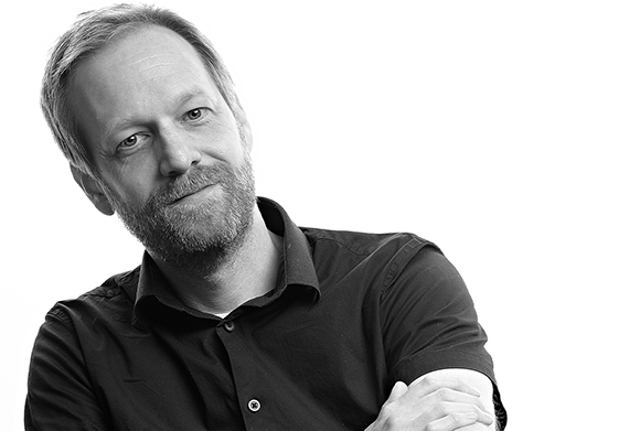 Black and white portrait of a man with a beard and short hair, tilting his head slightly and wearing a dark shirt.