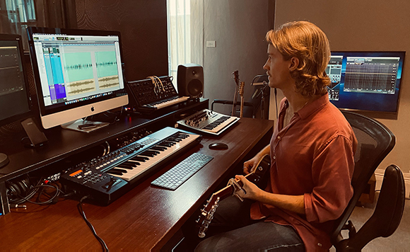 A man with wavy blonde hair sits at a desk playing an electric guitar in a music studio with multiple monitors.