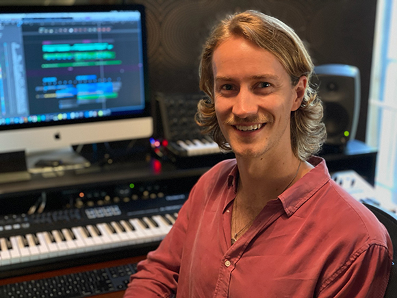 A man with blond hair and a mustache smiling in a music studio with a computer and keyboard.