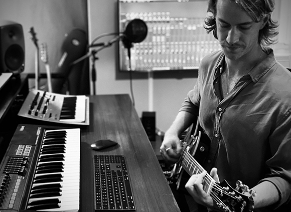 A man sits at a desk playing an electric guitar in a studio filled with musical equipment.