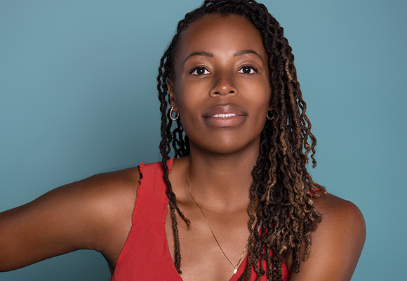 A woman with long dreadlocks and a red top looks forward against a blue background.