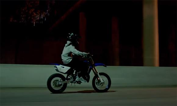 A person in a light grey hoodie and helmet riding a blue and white dirt bike on a road at night.
