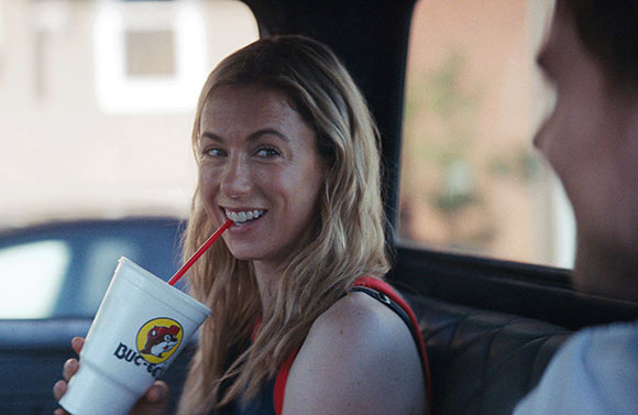 A blonde woman smiles while drinking from a white Buc-ee's cup in the backseat of a vehicle.
