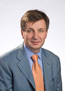 A man with short brown hair wearing a blue patterned suit, light blue shirt, and orange patterned tie.