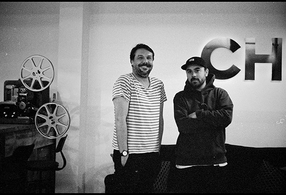 Black and white photo of two men standing indoors near a film projector and large letters on a wall.