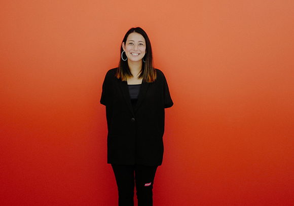 A smiling woman with long dark hair wearing a black blazer and black pants against a bright orange background.