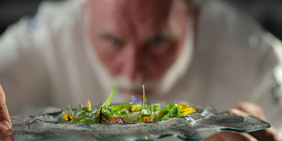 A blurred chef looks closely at a colorful, garnished dish on a clear glass plate.