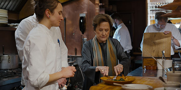 A woman in a dark scarf inspects a plate of food while kitchen staff in white uniforms work in the background.