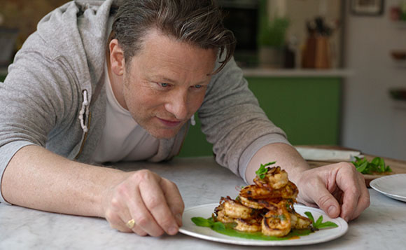 A man in a grey hoodie leans over a white plate of grilled shrimp garnished with green herbs.