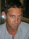 A man with short dark hair wearing a light gray button-down shirt looking forward.