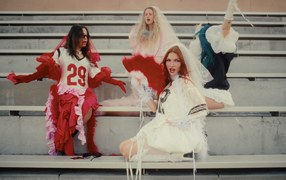 Four models in eclectic, ruffled, and voluminous outfits pose on outdoor concrete stadium seating.