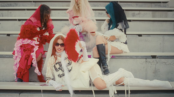 Four people in voluminous, textured, and colorful avant-garde clothing sit on concrete stairs.