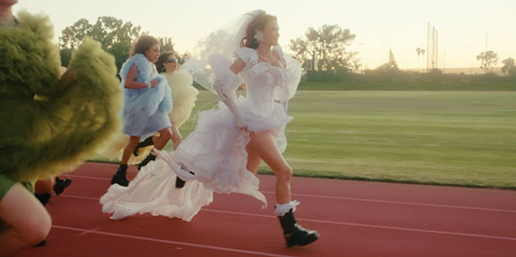 People in large, fluffy, colorful costumes running on a red athletic track during sunset.