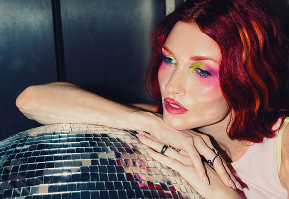 A person with wavy red hair and colorful makeup leans their chin on a large disco ball.