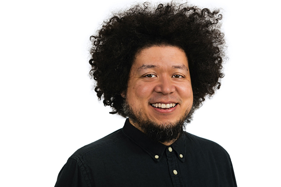 A smiling man with voluminous dark curly hair and a dark beard wearing a black button-down shirt.