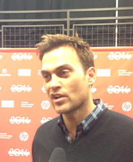 A man with short brown hair and a plaid shirt under a dark sweater speaks into a microphone. A red backdrop with '2014' is be