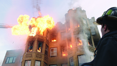 A firefighter in gear watches a multi-story brick building erupt in large orange flames and smoke.