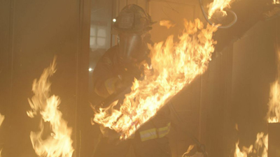 A firefighter in full protective gear stands amidst large, bright orange flames.