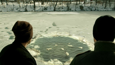 Two people in winter clothing look out over a frozen body of water with a large circular hole in the ice.
