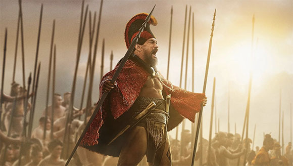 A shirtless warrior in a red cape and helmet shouts while holding a spear amidst a crowd of soldiers.