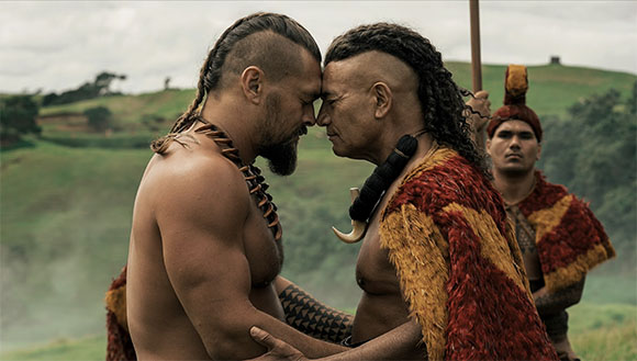Two shirtless men with traditional hairstyles and tattoos press their foreheads together outdoors.